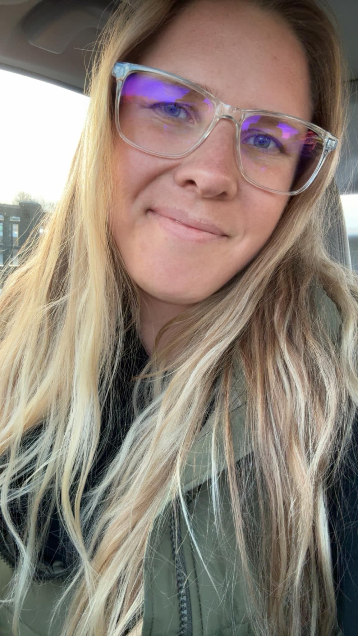 A smiling blonde woman wearing clear-framed glasses with blue tint takes a selfie in a car.
