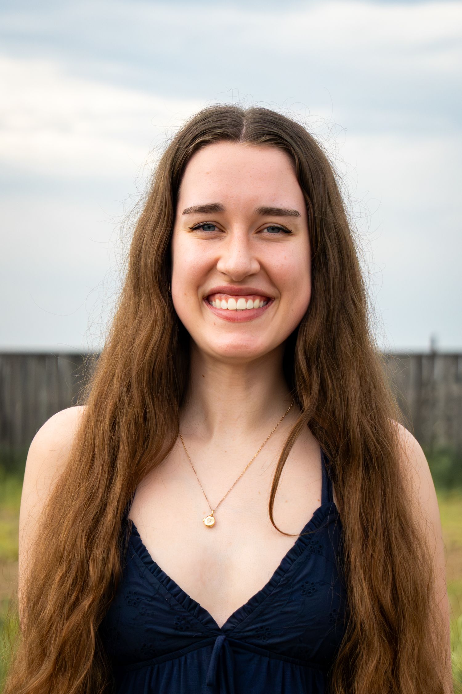 Smiling young woman with long brown hair wearing a navy dress and gold necklace outdoors.