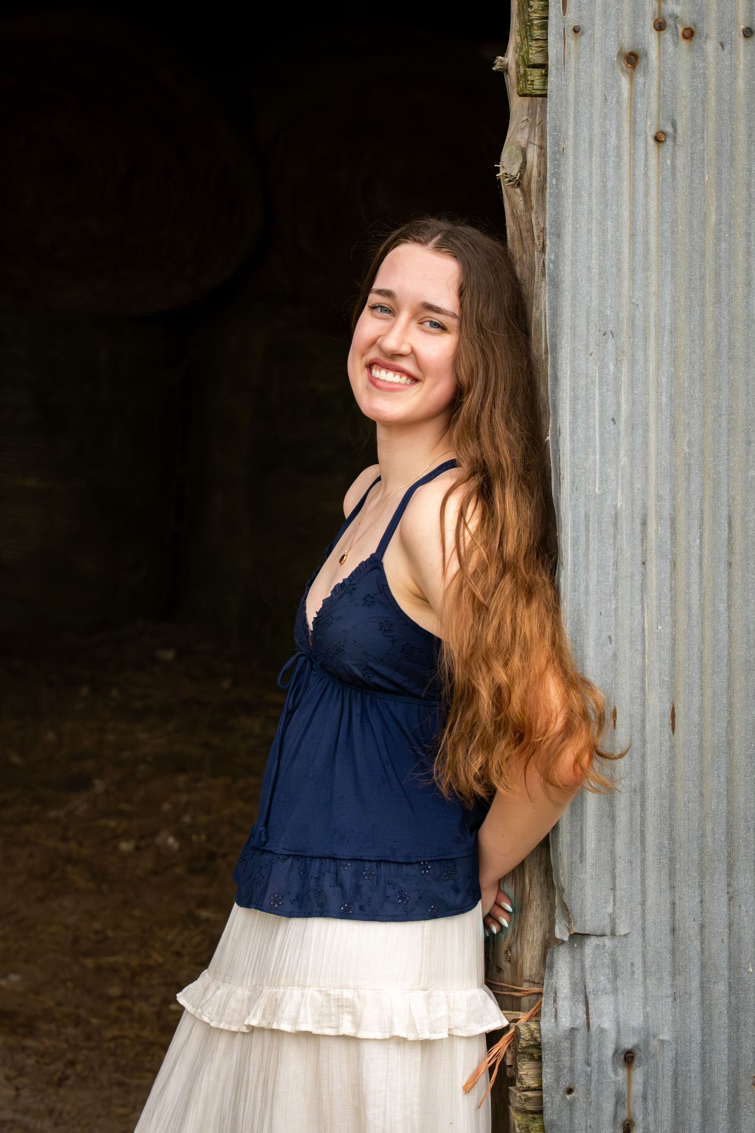 A cheerful young woman with long hair wearing a navy top and white skirt posing by a rustic barn door.