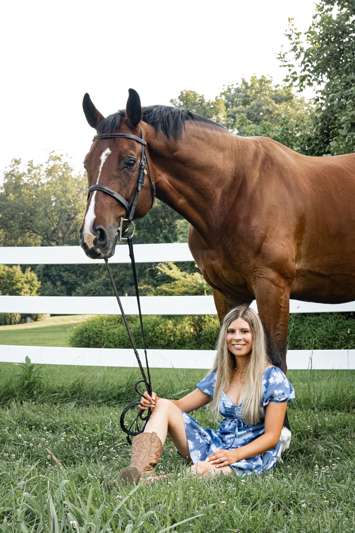 Young woman in blue floral dress sitting on grass beside a bay horse at a white fence farm.