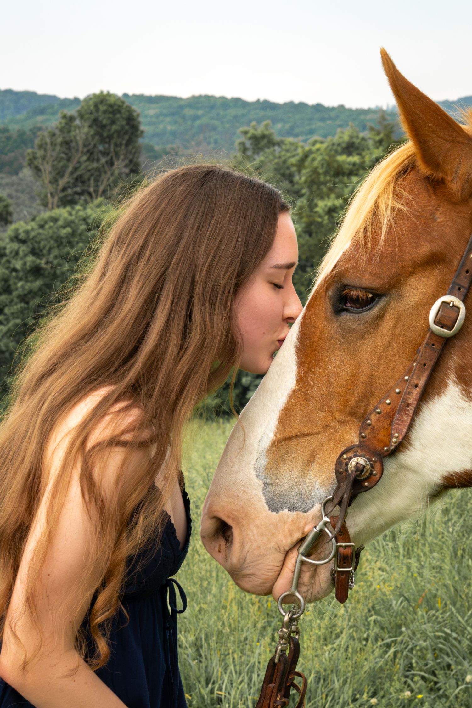 Young woman with long brown hair gently kissing a chestnut paint horse on the nose in a green field.