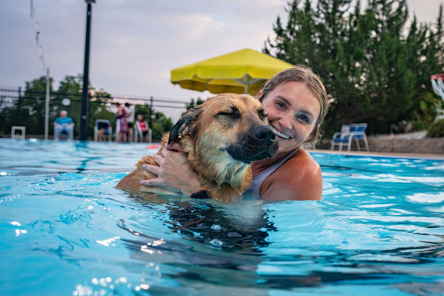 A happy woman embracing her large wet dog while swimming in an outdoor swimming pool.