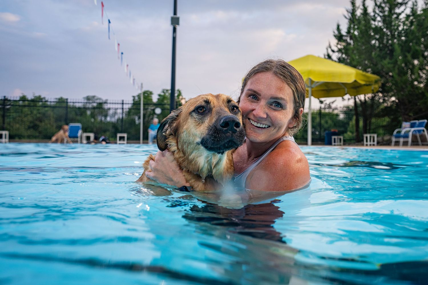 A smiling woman hugging her wet dog while swimming together in an outdoor pool.