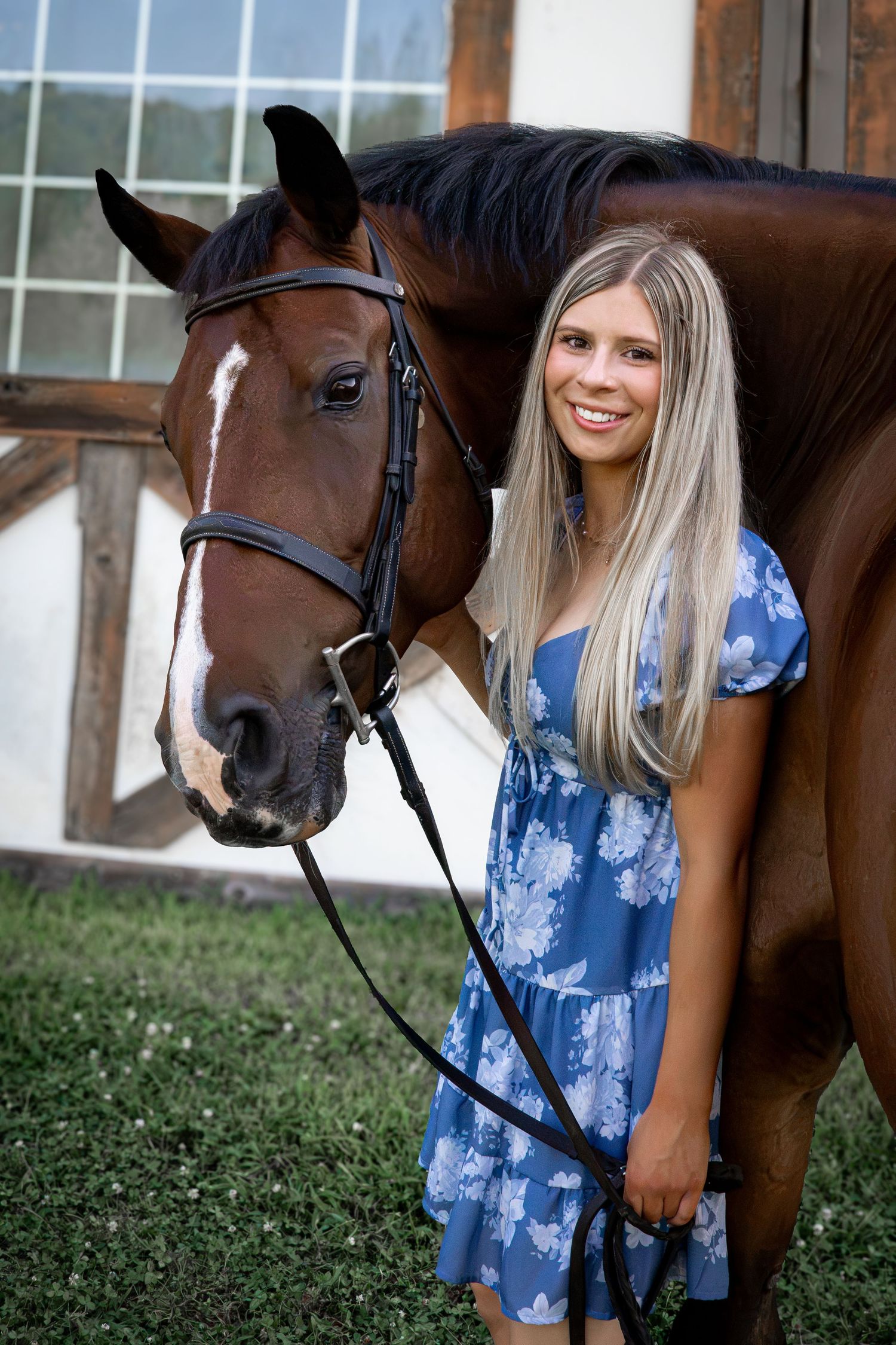 Young blonde woman in blue floral dress smiling beside a brown horse with white blaze, held by black bridle outdoors.