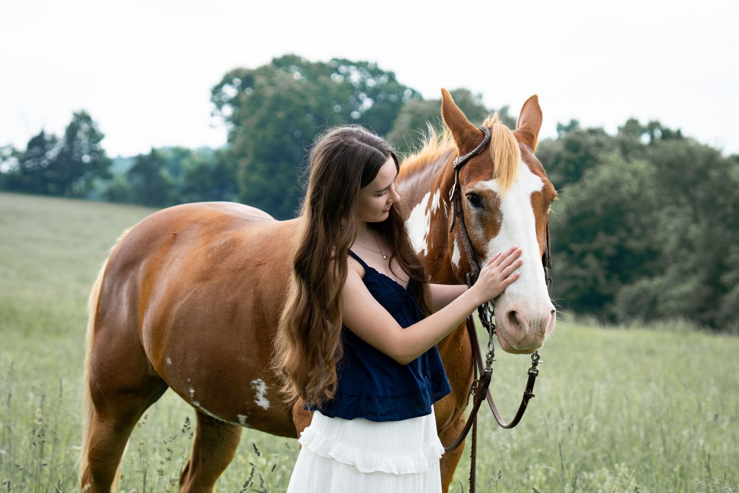 A young woman with long brown hair gently touching the face of a chestnut Paint horse in a field.