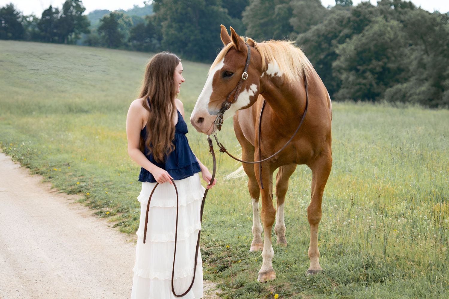 Young woman in navy top and white skirt standing beside a chestnut horse on a scenic country road.