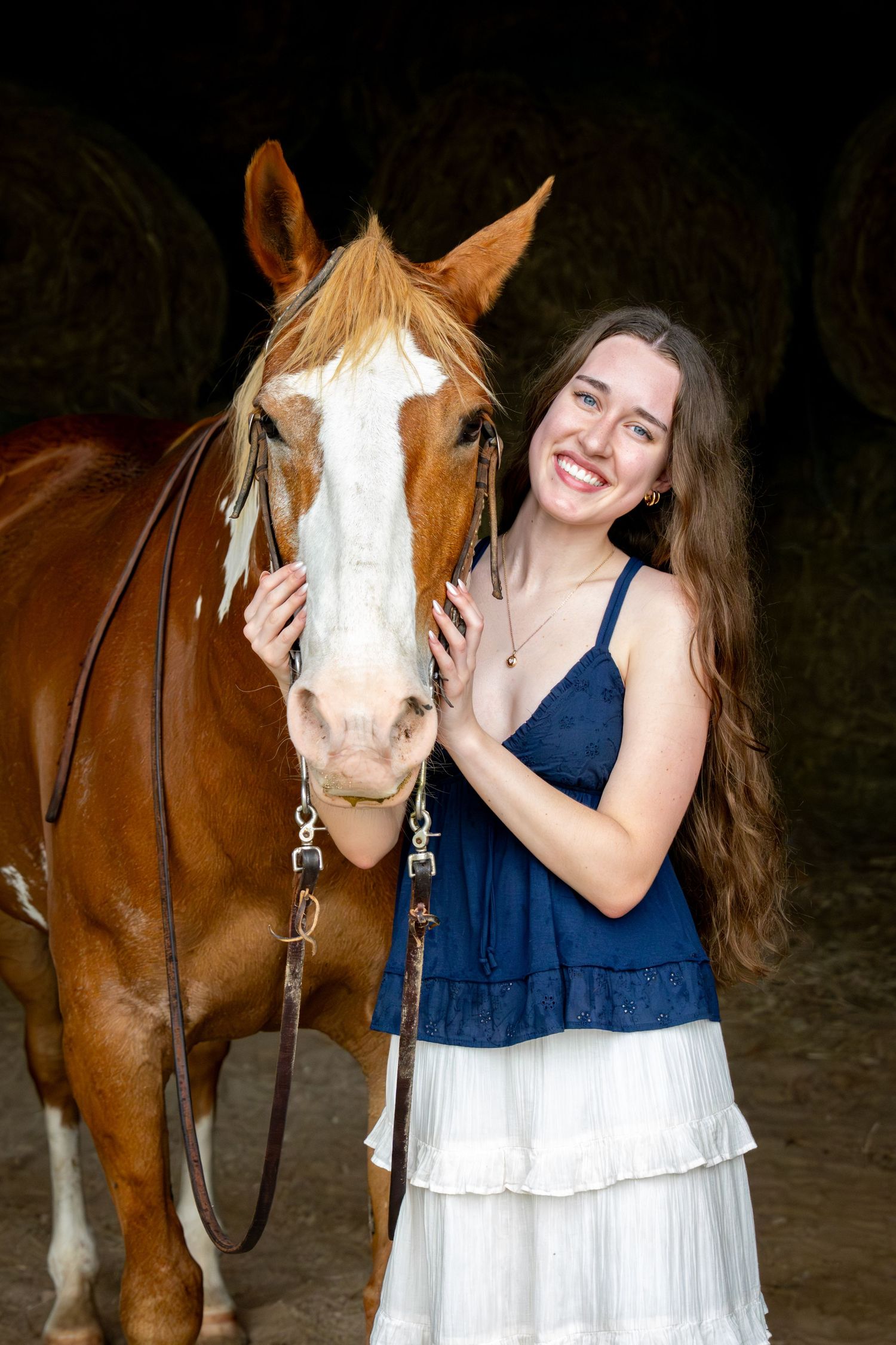 Happy young woman with long brown hair in blue top smiling beside a chestnut horse in a barn.