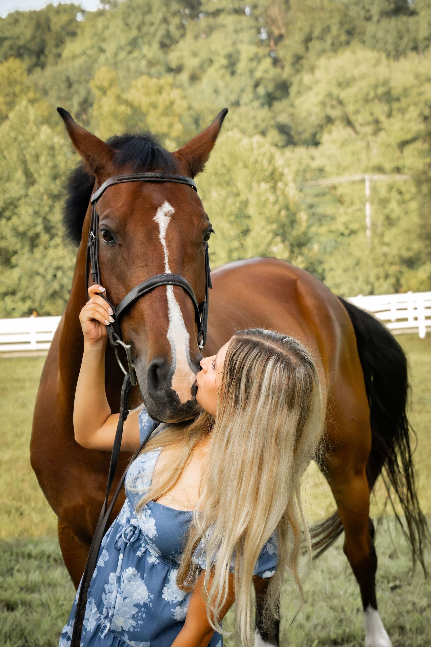 Blonde woman in blue floral dress kissing a brown horse with white blaze, standing by white fence.