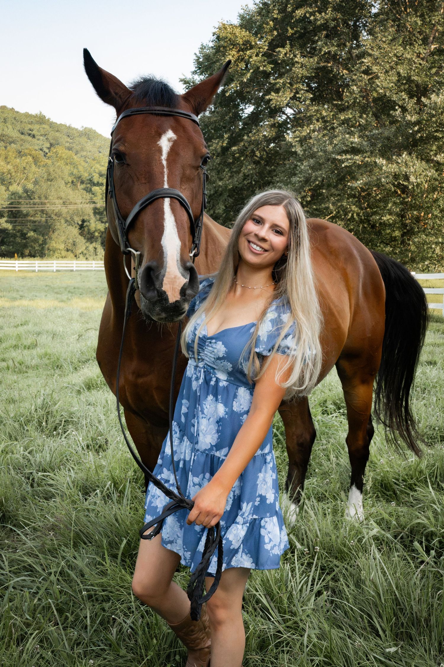 Young blonde woman in blue floral dress smiling beside a chestnut horse in a lush green pasture.