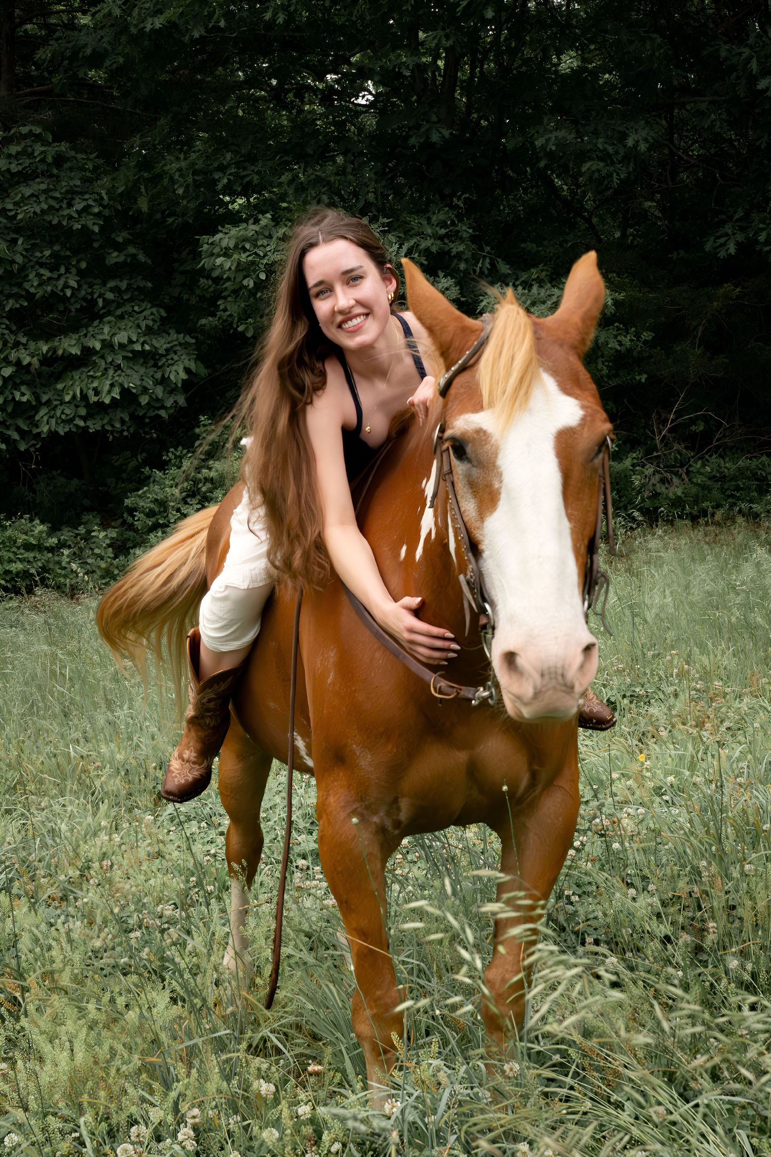 Smiling young woman with long hair riding a chestnut paint horse in a lush green meadow.