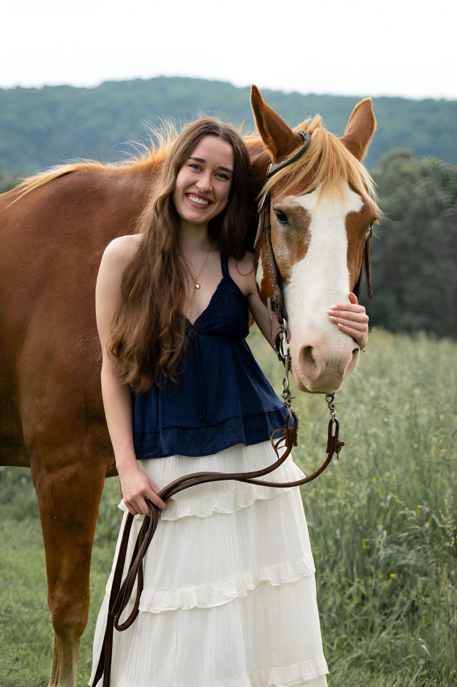 A smiling young woman in a navy dress and white skirt posing beside a chestnut Paint horse in a field.