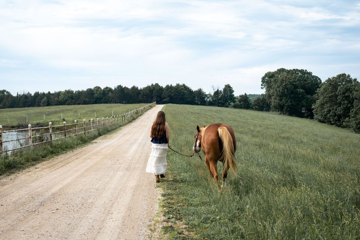 Young woman in white skirt leading a chestnut horse along a rural dirt road through green fields.