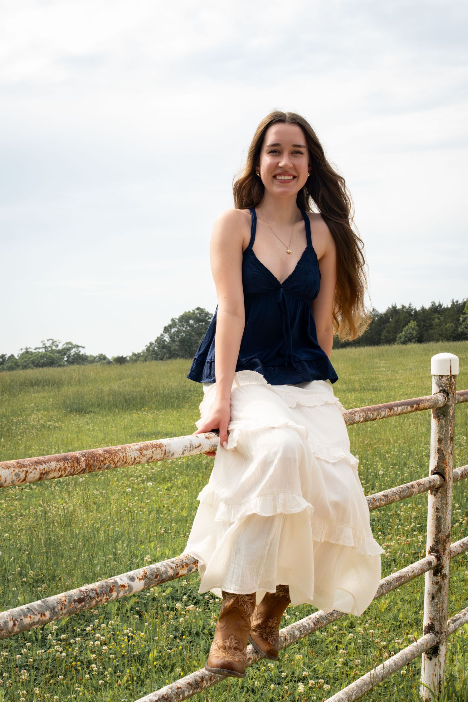 Smiling young woman in navy top and white ruffled skirt leaning on a rustic fence in a meadow.
