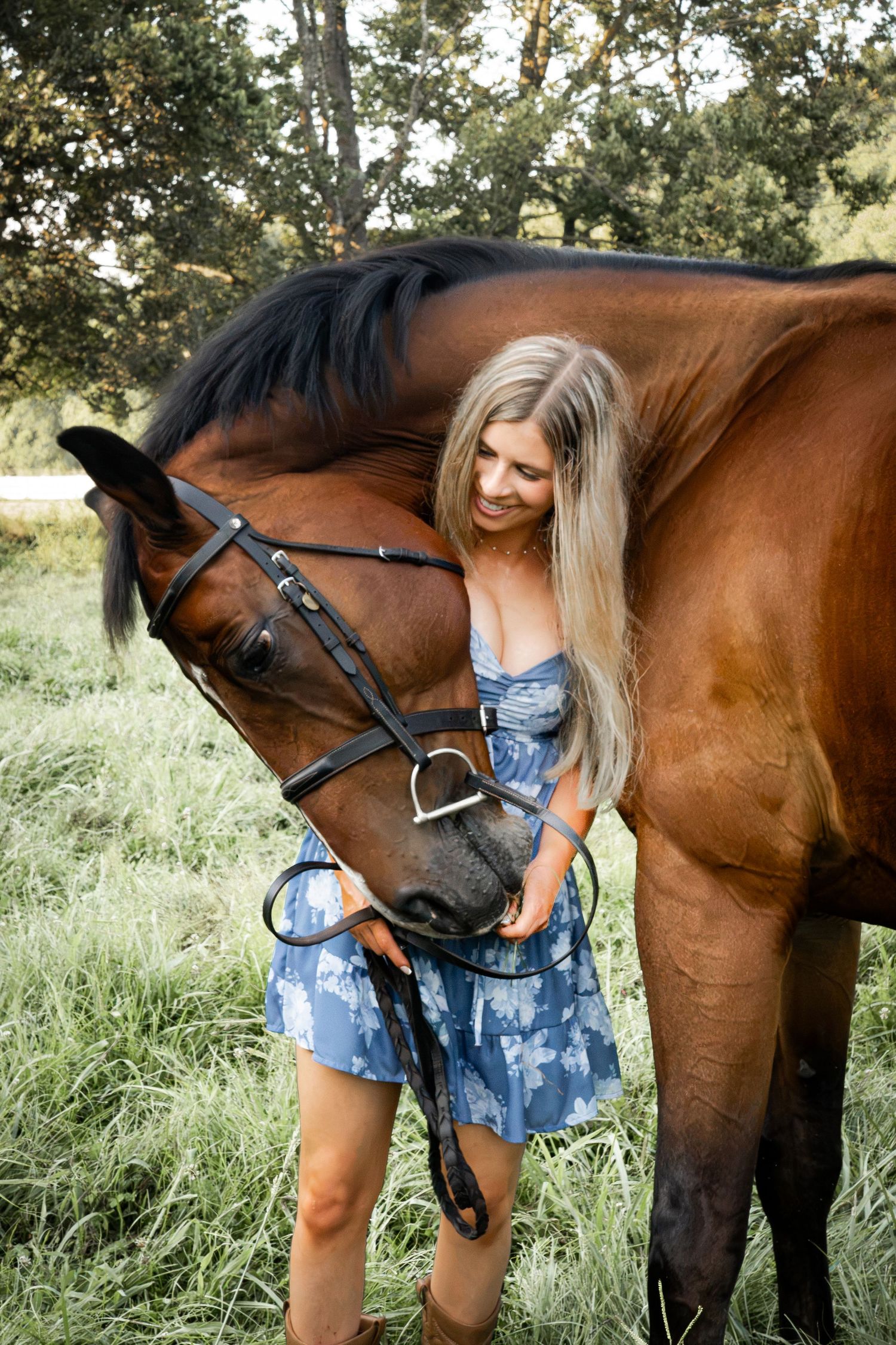 Blonde woman in blue floral dress tenderly interacting with a bay horse wearing a bridle in a green field.