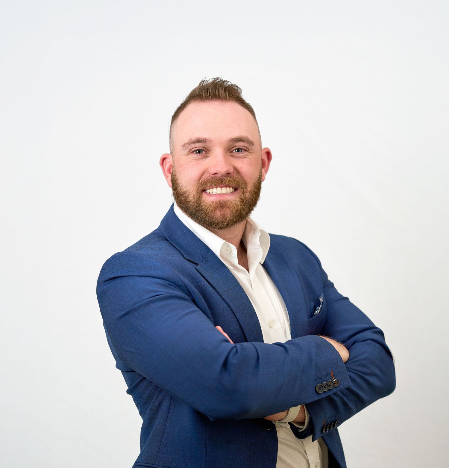 Confident bearded man in blue suit jacket posing with arms crossed on white background.