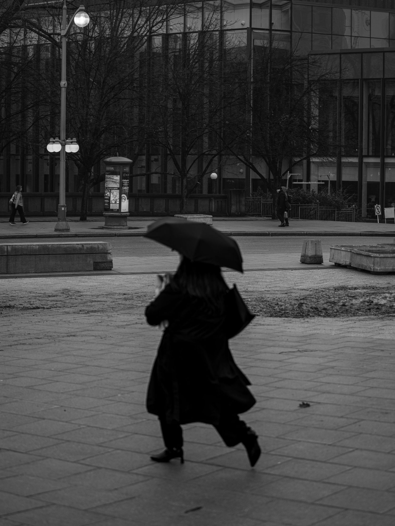 Black and white photo of person walking with umbrella on rainy urban plaza.