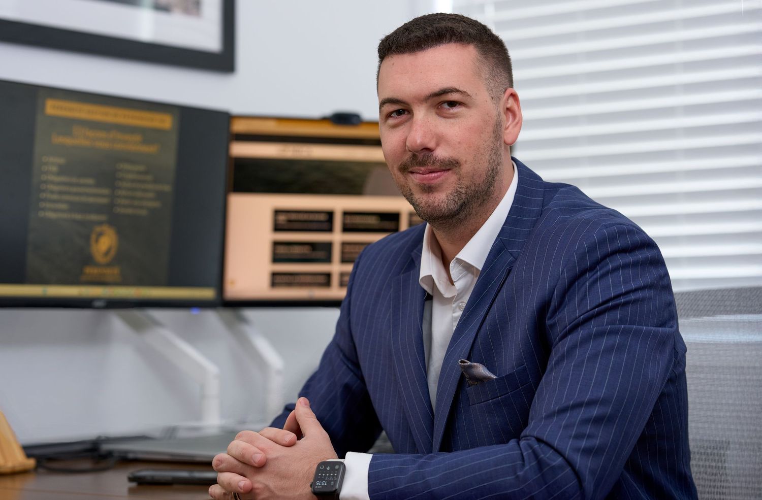 Professional man in blue pinstripe suit sitting at desk with monitors in modern office.