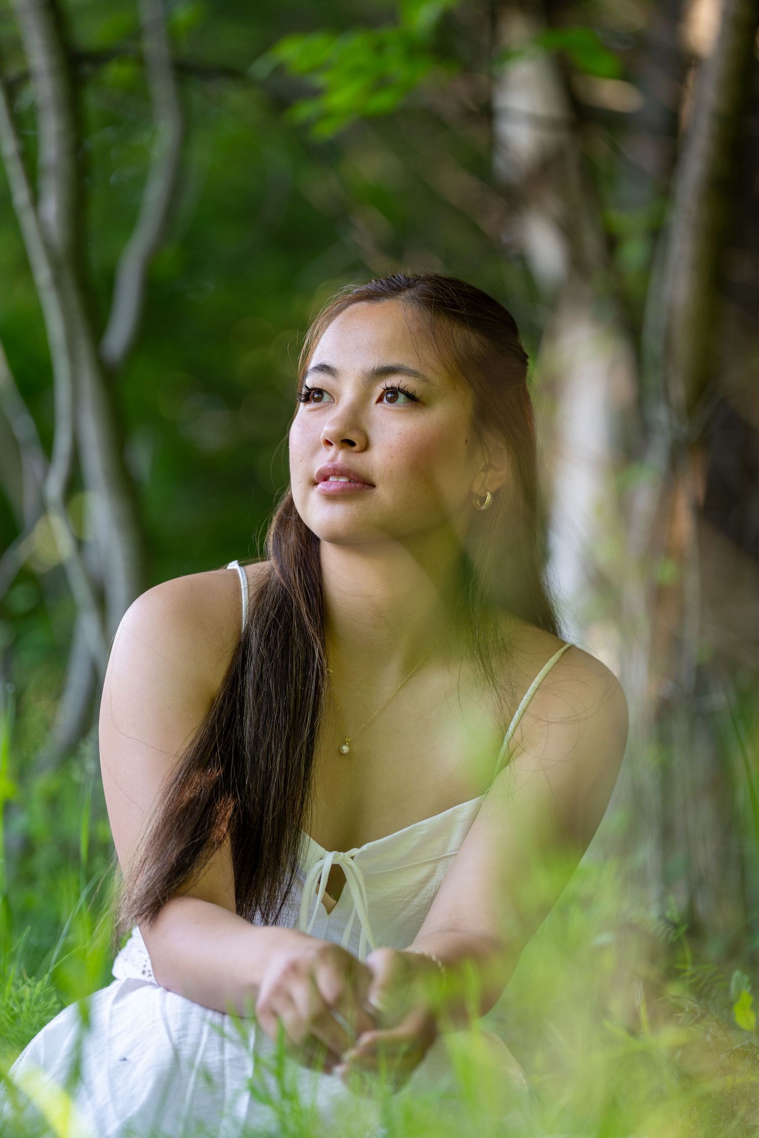 Young woman in white dress sitting on grass gazing upward in a serene forest setting.