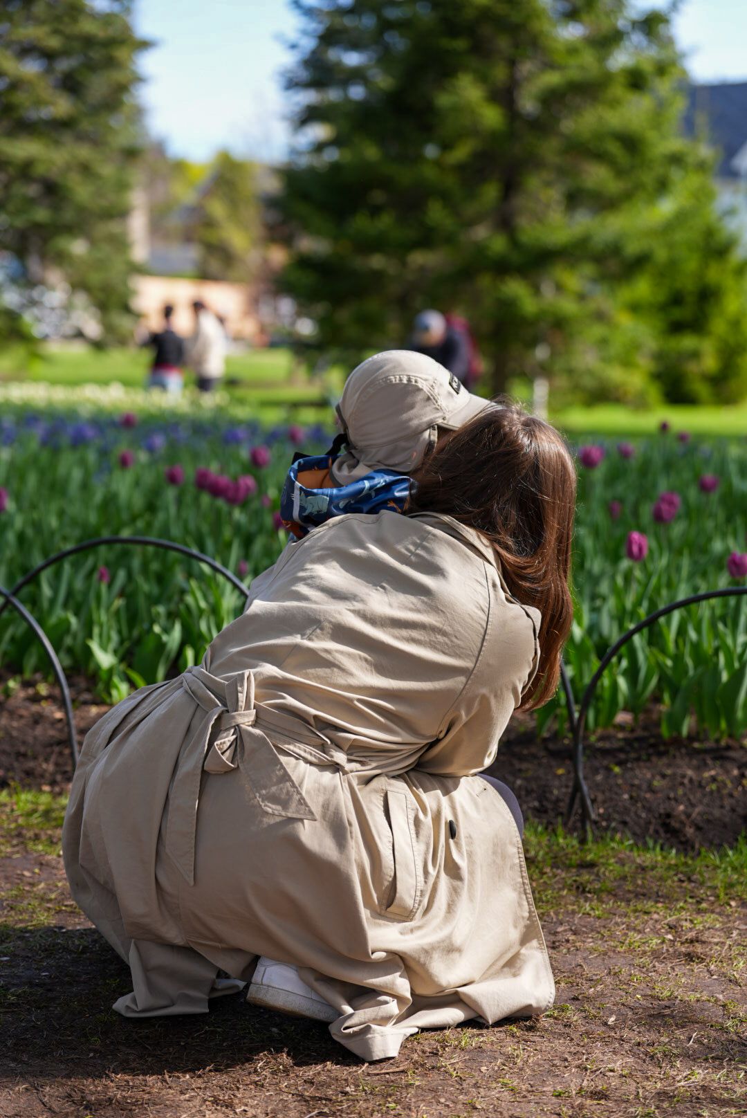 Woman in beige trench coat crouching to photograph colorful tulips in a garden.
