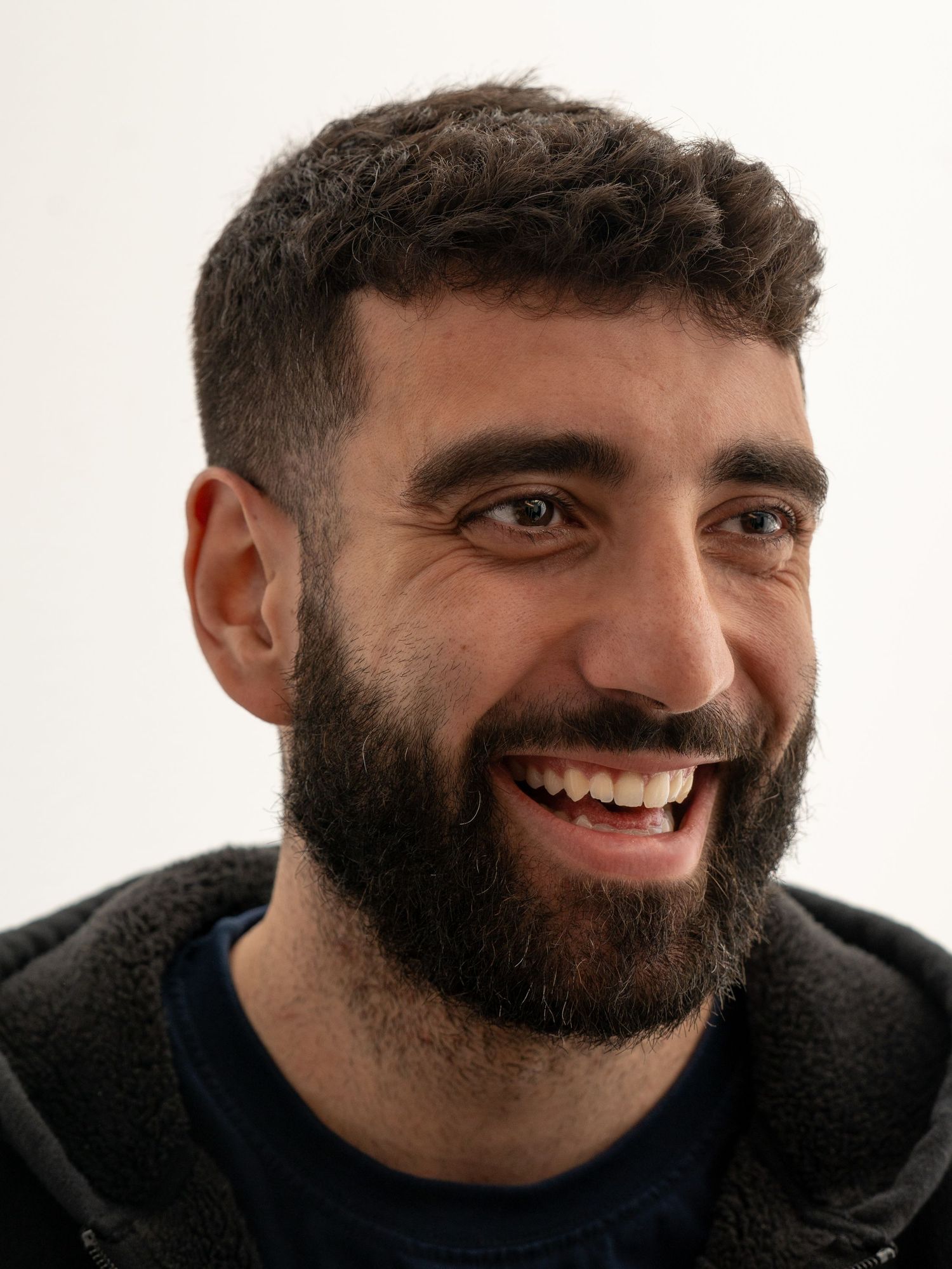 Close-up portrait of a smiling bearded man with curly hair wearing a dark jacket against white background.