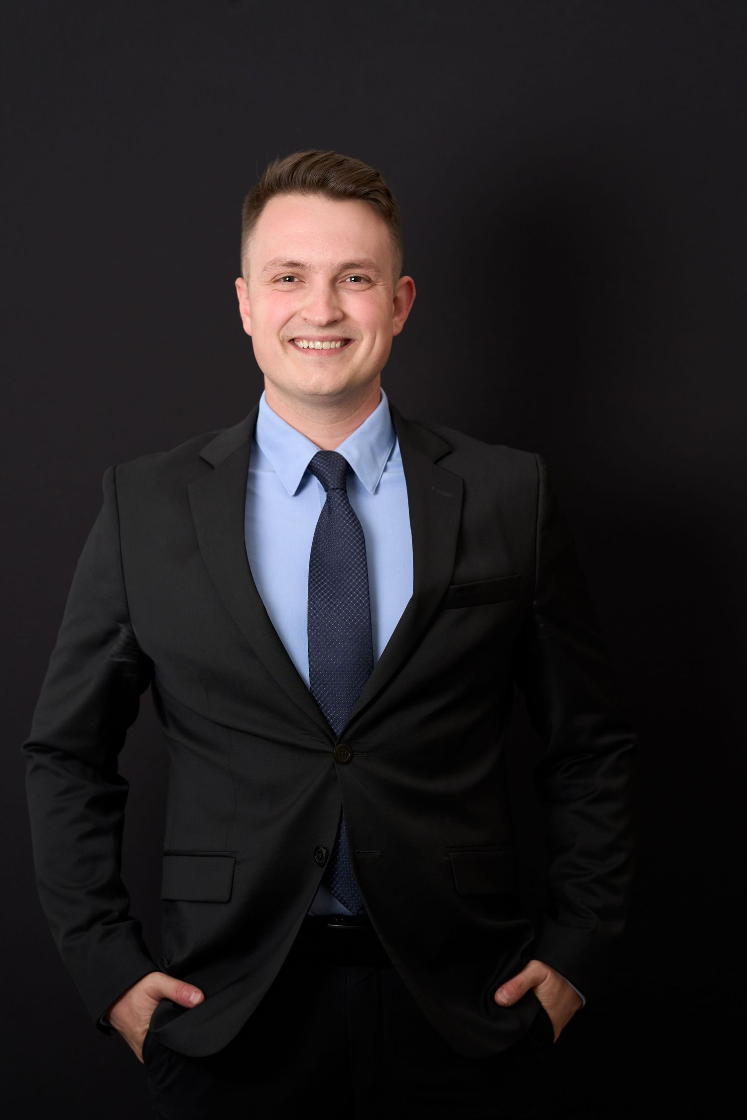 Smiling young man in black suit and blue shirt standing confidently against dark background.