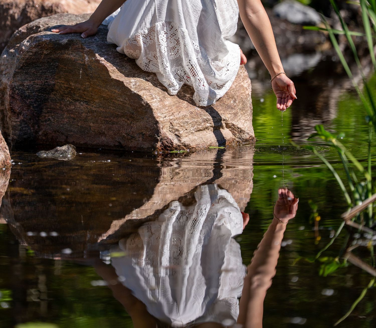 A woman in a white dress sitting on a rock with her reflection mirrored in calm water below.