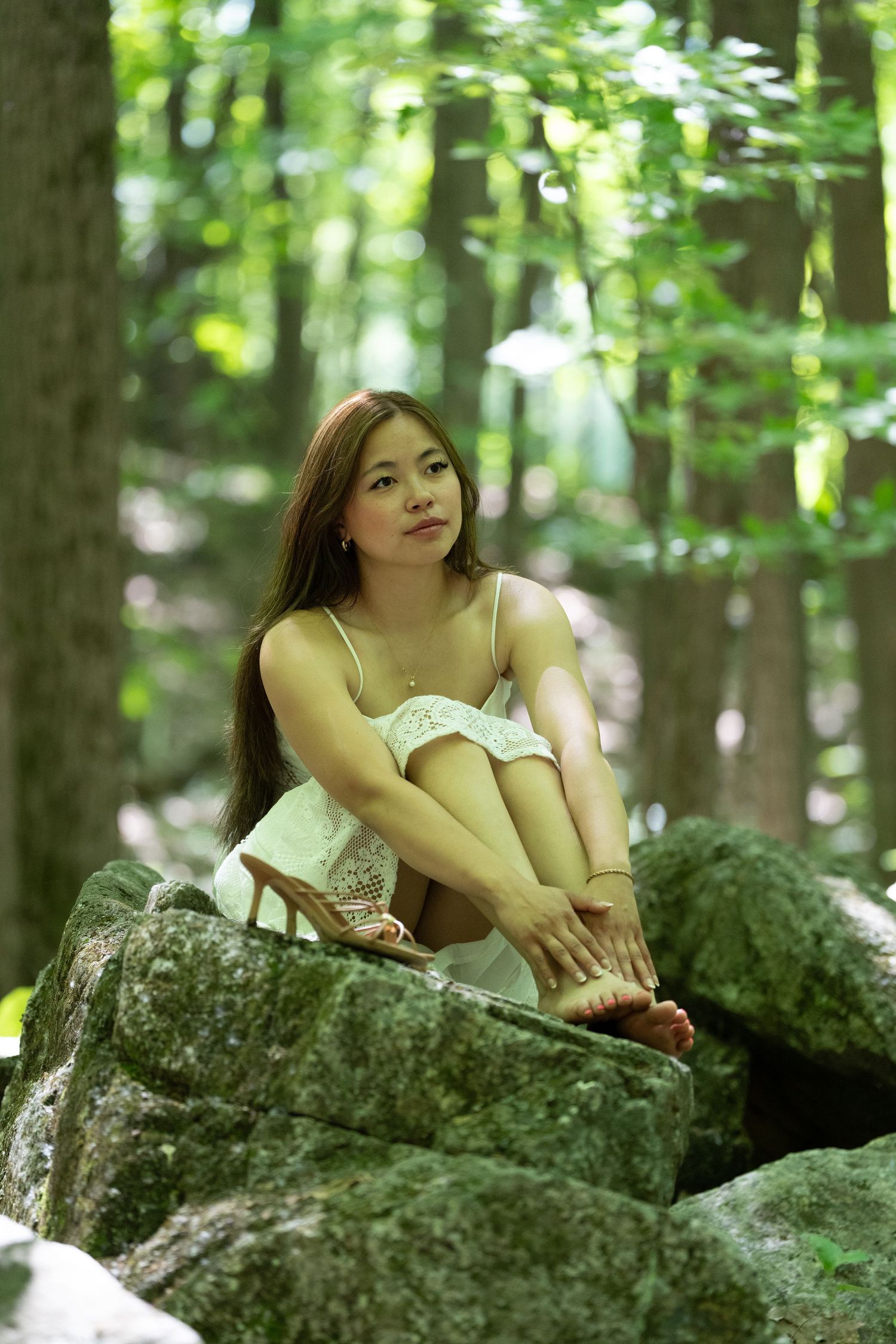Young Asian woman in white dress sitting on mossy rocks in a green forest.