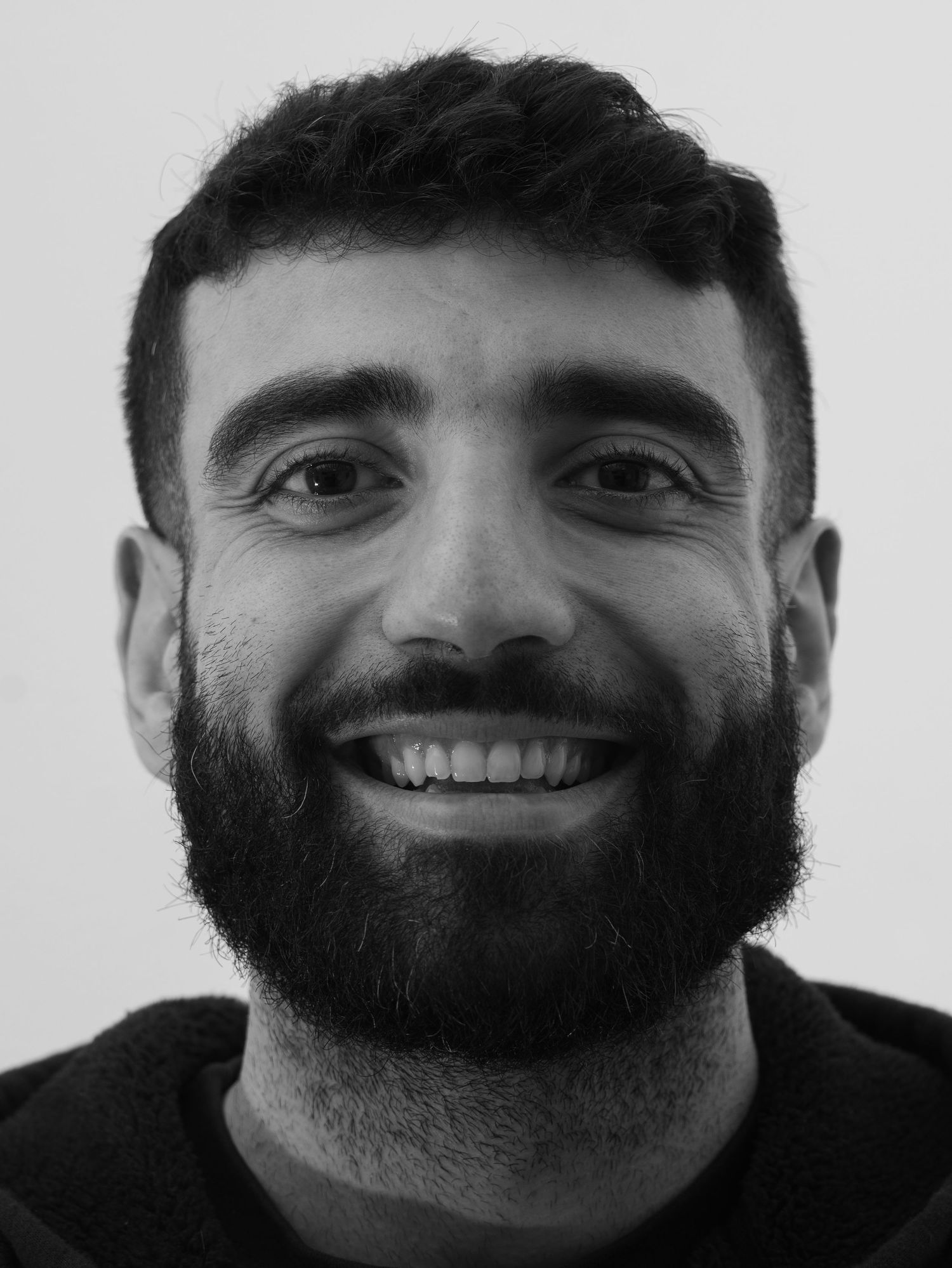 Black and white close-up portrait of a smiling bearded man with short dark hair against light background.