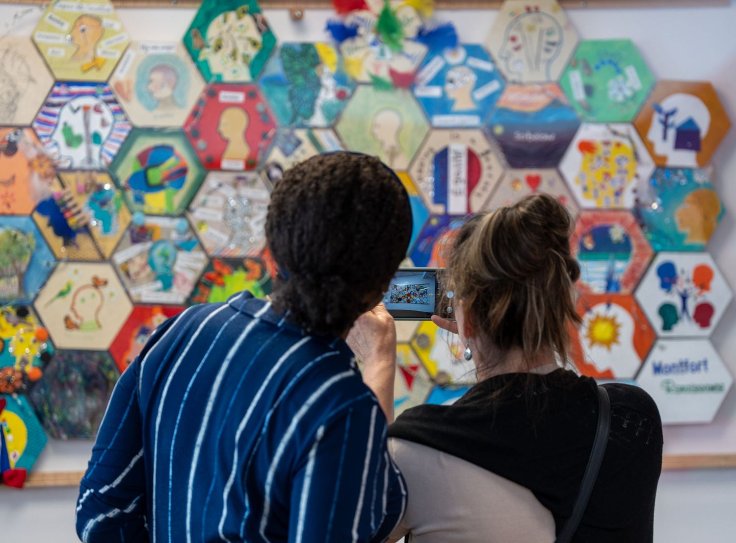 Two visitors photograph a colorful hexagonal mosaic wall display at an art exhibition.