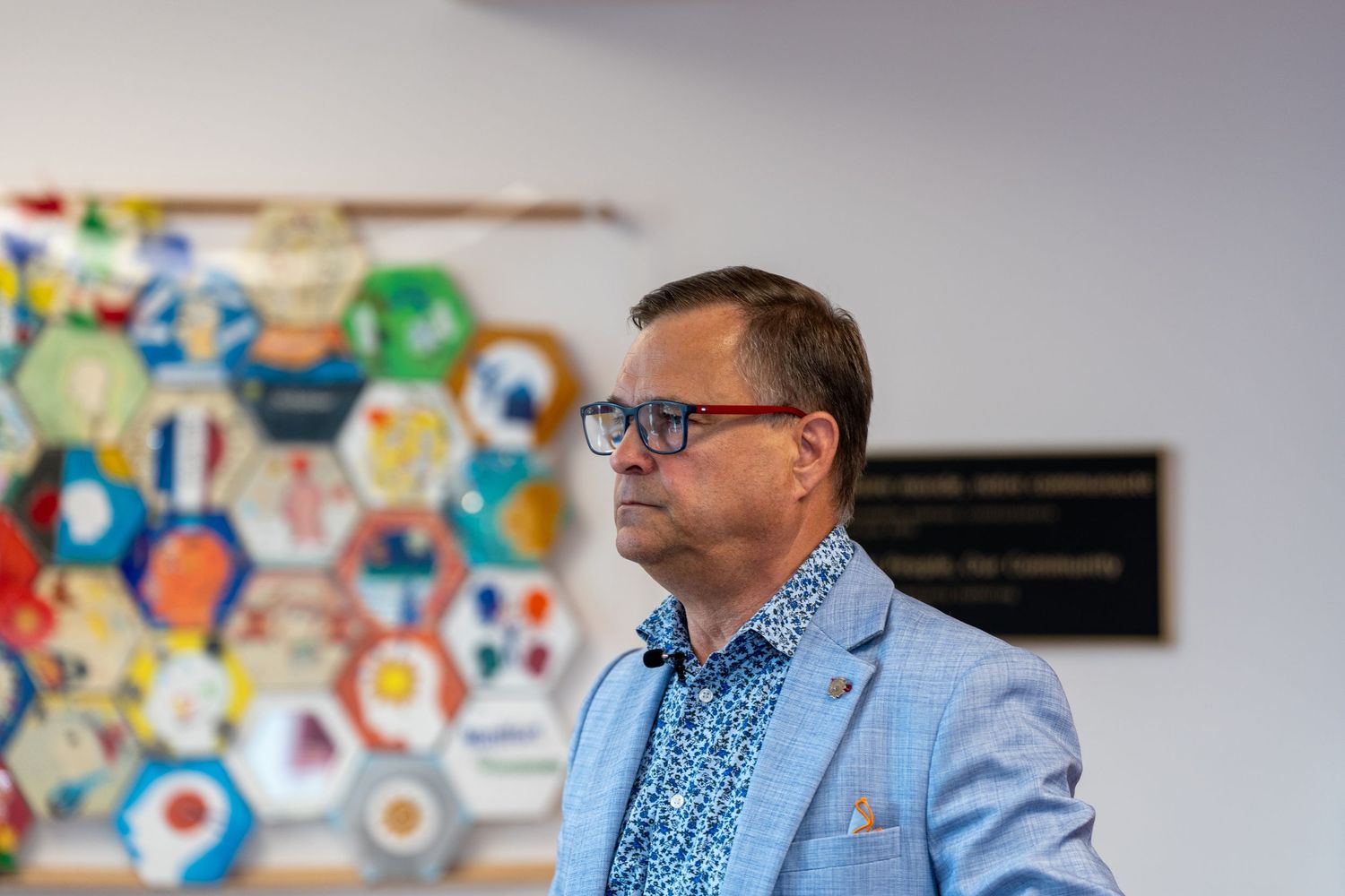 Man in blue blazer and glasses stands before colorful hexagonal art display at indoor event.