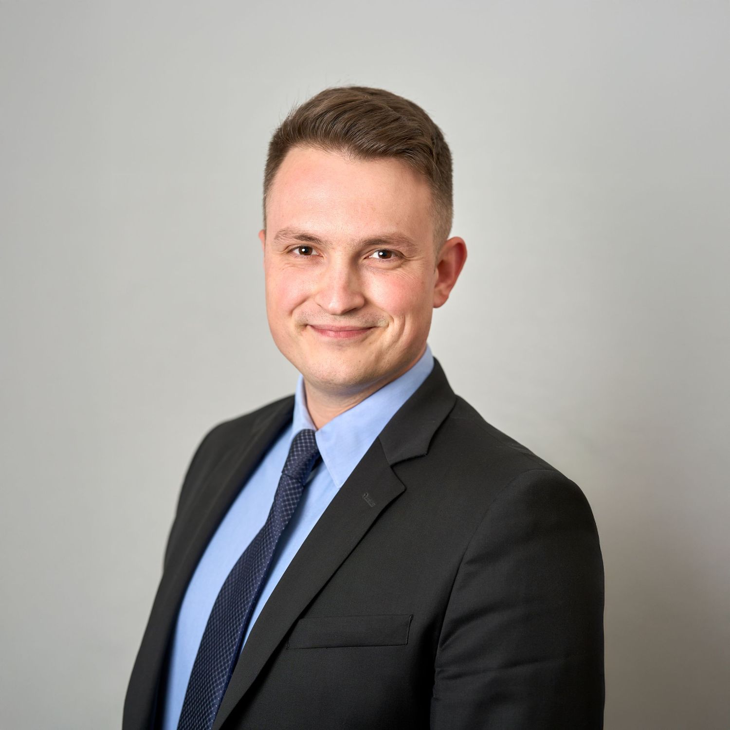 Professional male headshot of young man in dark blazer and blue shirt with navy tie.