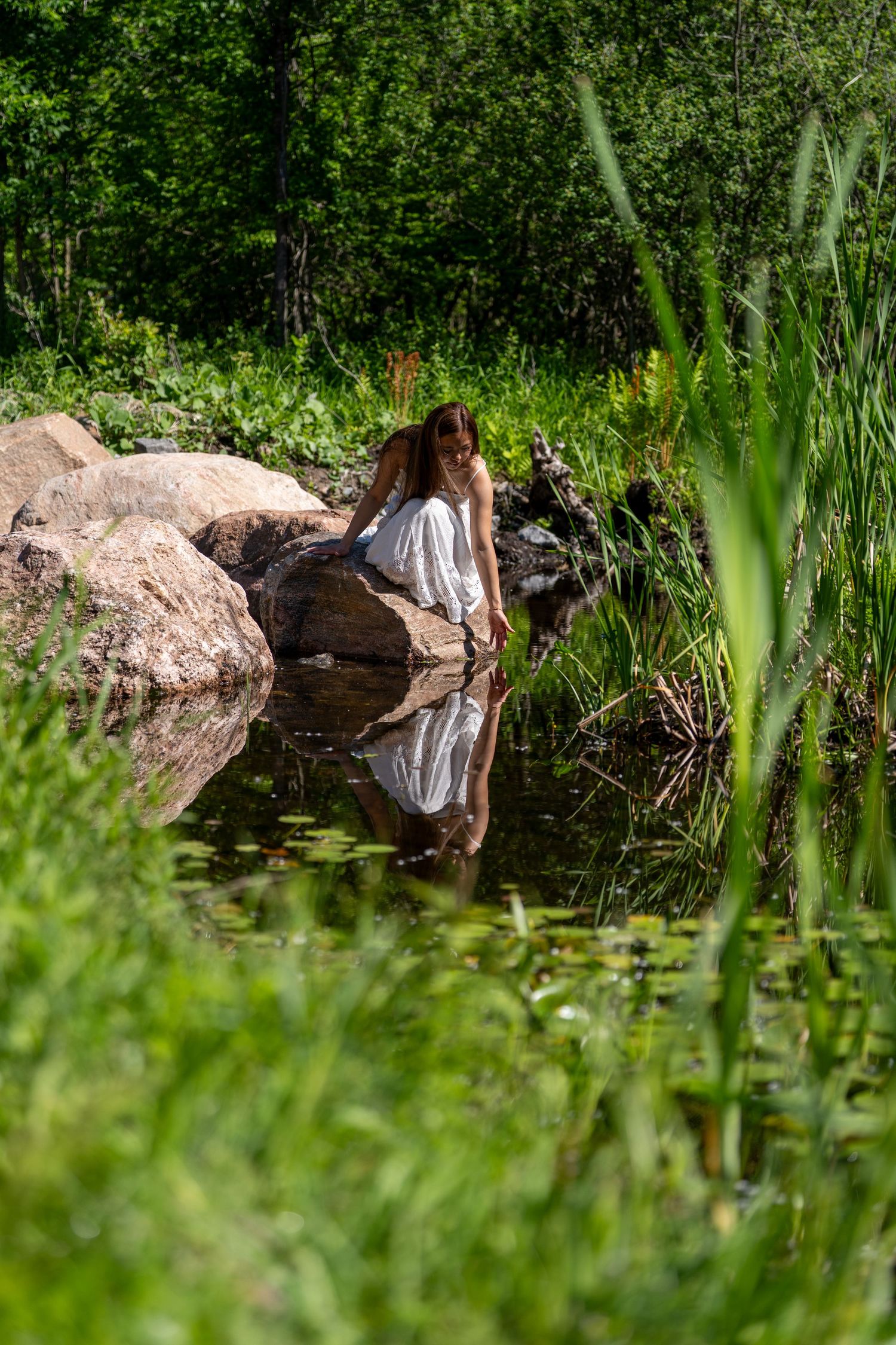A young woman in a white dress sitting on rocks beside a tranquil pond surrounded by lush greenery.