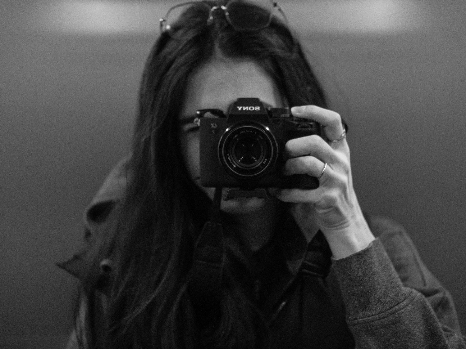 Young woman with long dark hair taking a mirror selfie while holding a Sony camera in black and white.