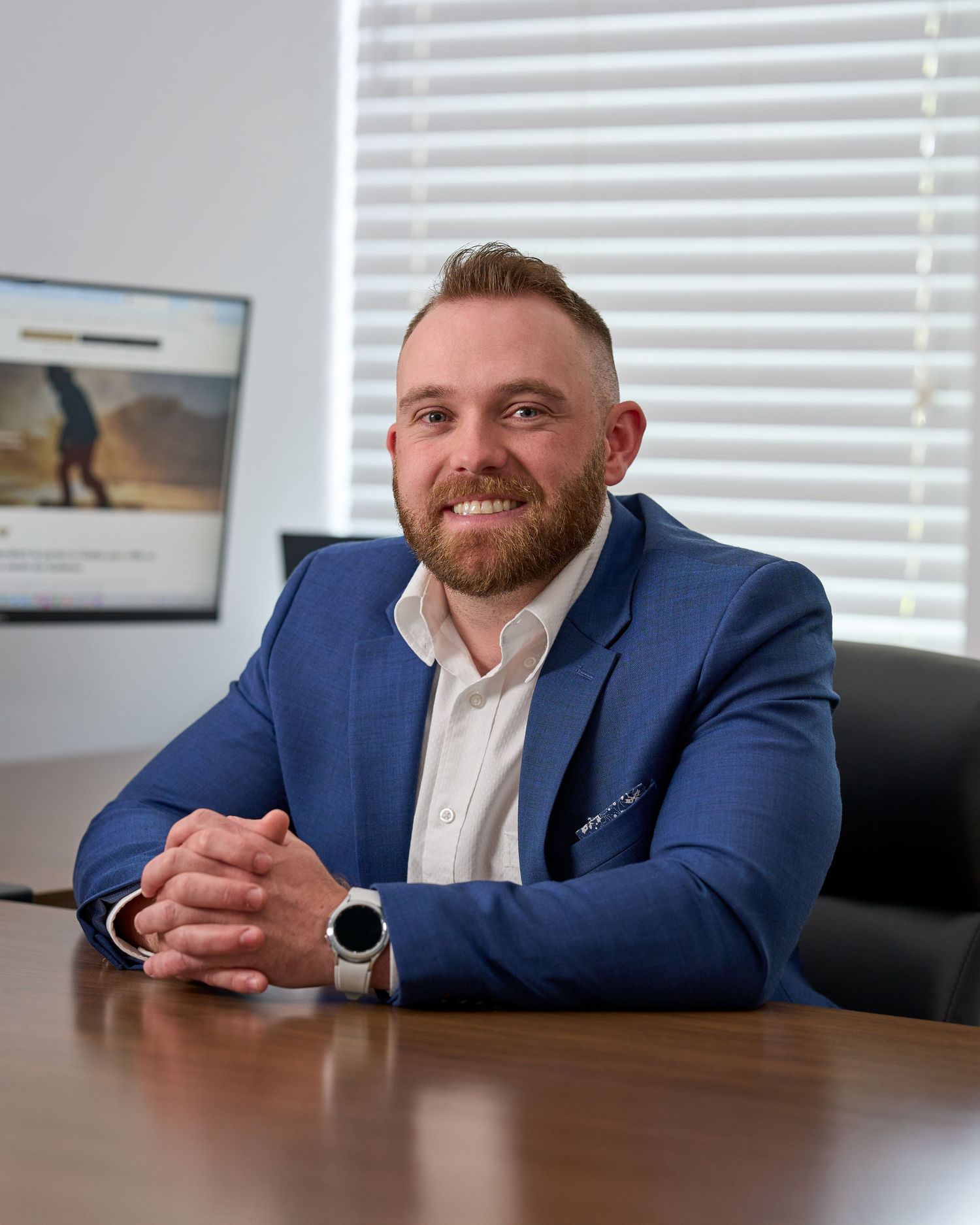Smiling bearded man in blue blazer seated at conference table in bright modern office.