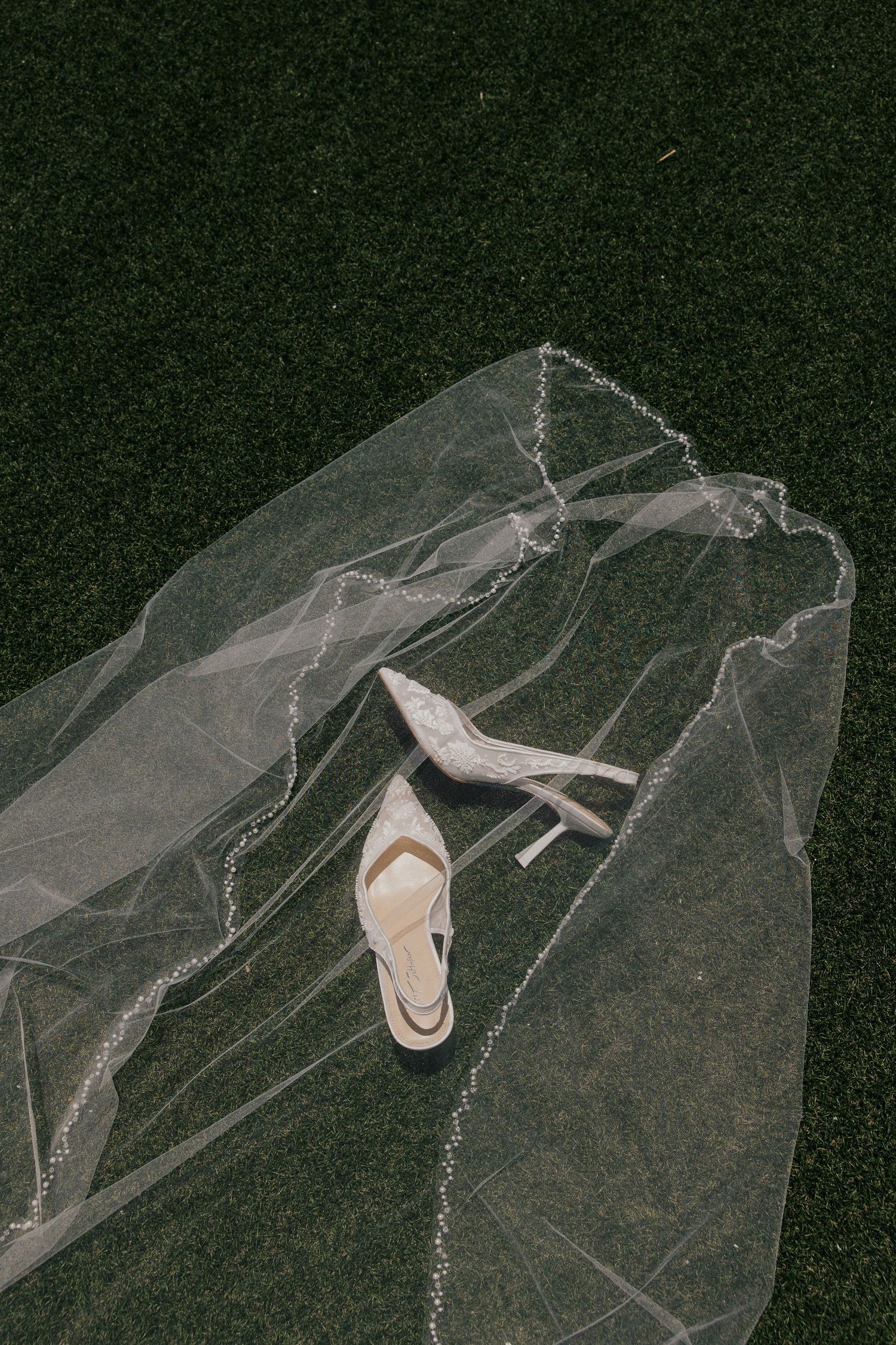 Bridal heels resting on a sheer wedding veil with crystal trim, laid out on lush green grass.