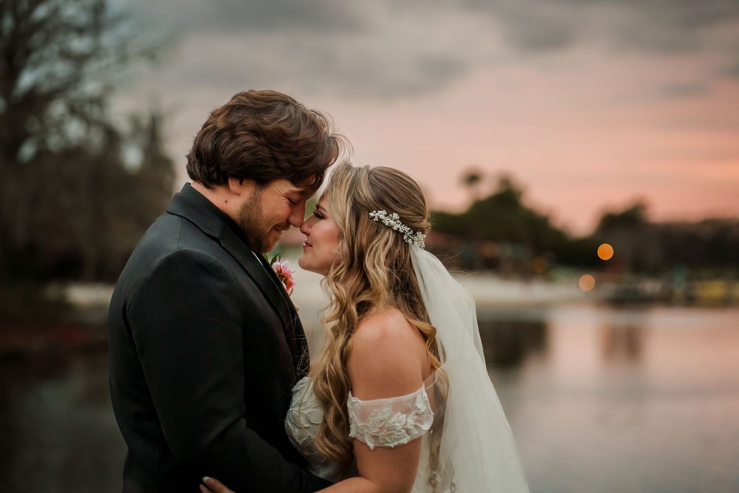 Bride and groom share a romantic moment near a lake at sunset, with bride in lace gown and veil.