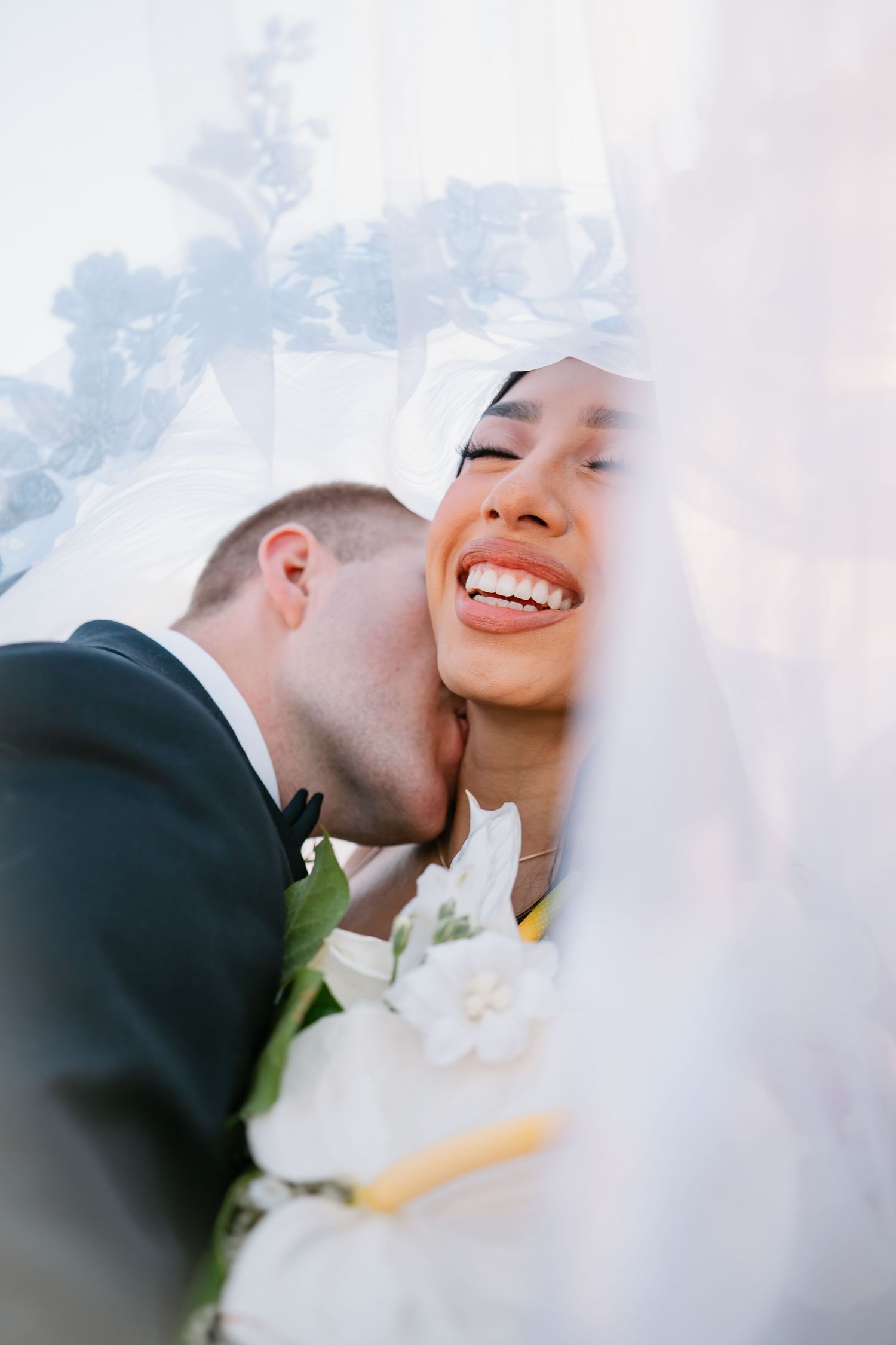 Groom kissing bride on cheek under wedding veil, bride laughing joyfully holding white bouquet.