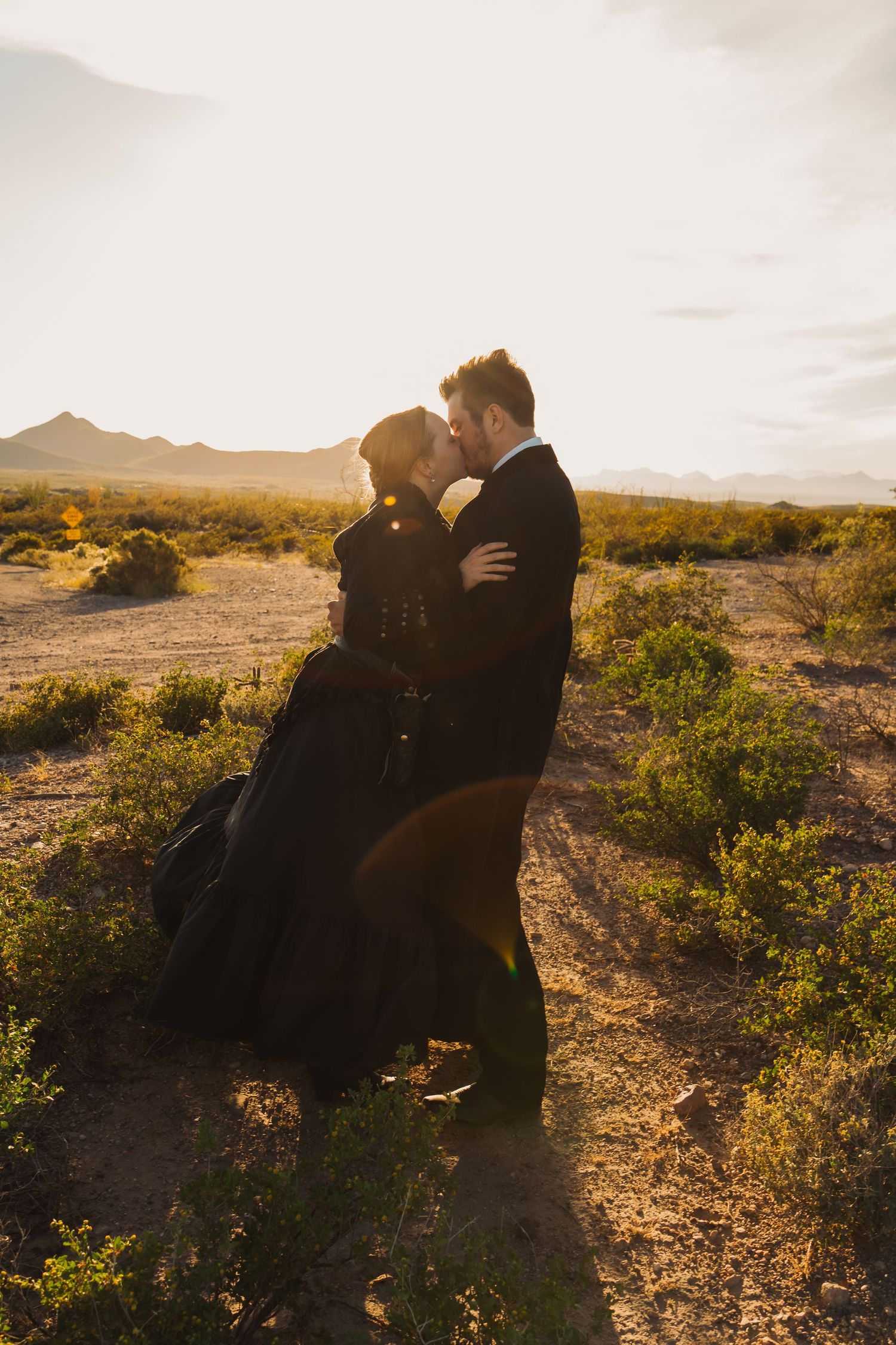 Couple in black formal wear sharing a romantic kiss at golden sunset in a desert landscape with mountains in the background.