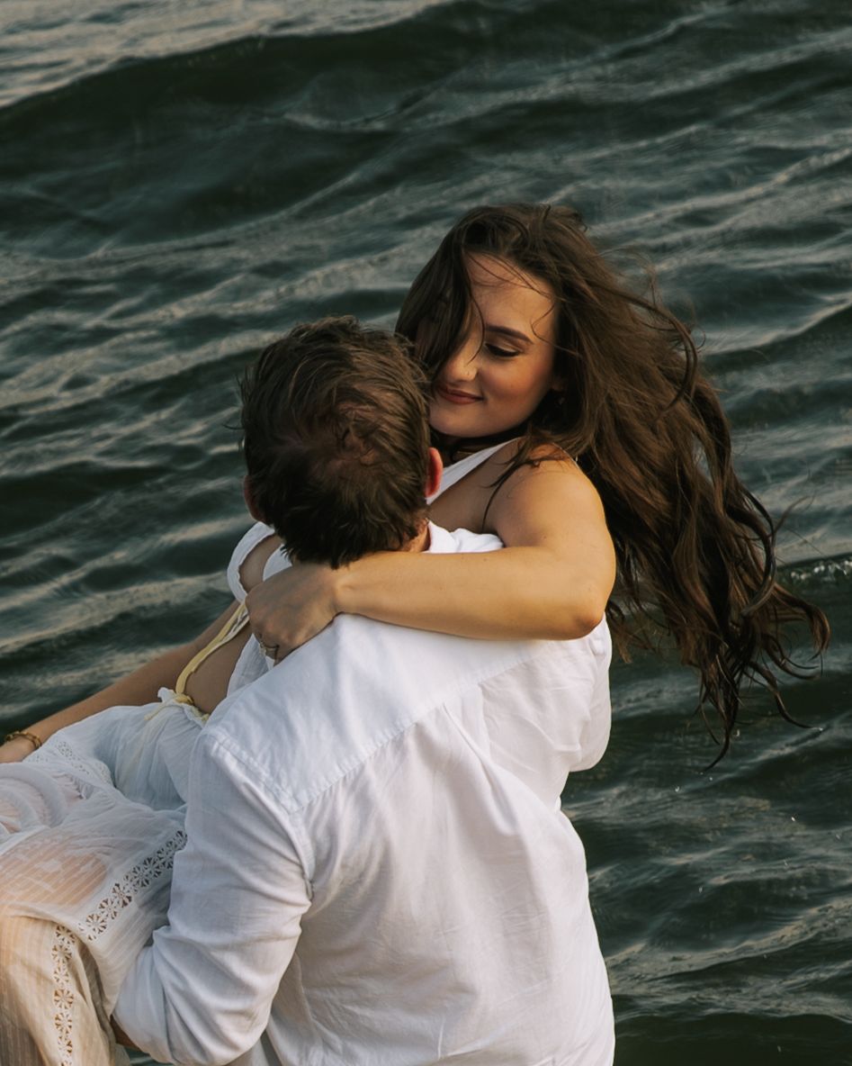 Couple embracing romantically by the ocean, woman with long dark hair smiling as man lifts her near water.