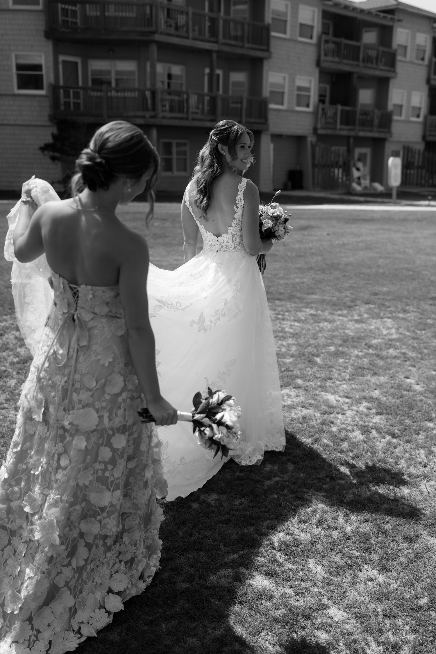 Black and white photo of bride in lace gown holding bouquet, assisted by bridesmaid in floral dress outdoors.