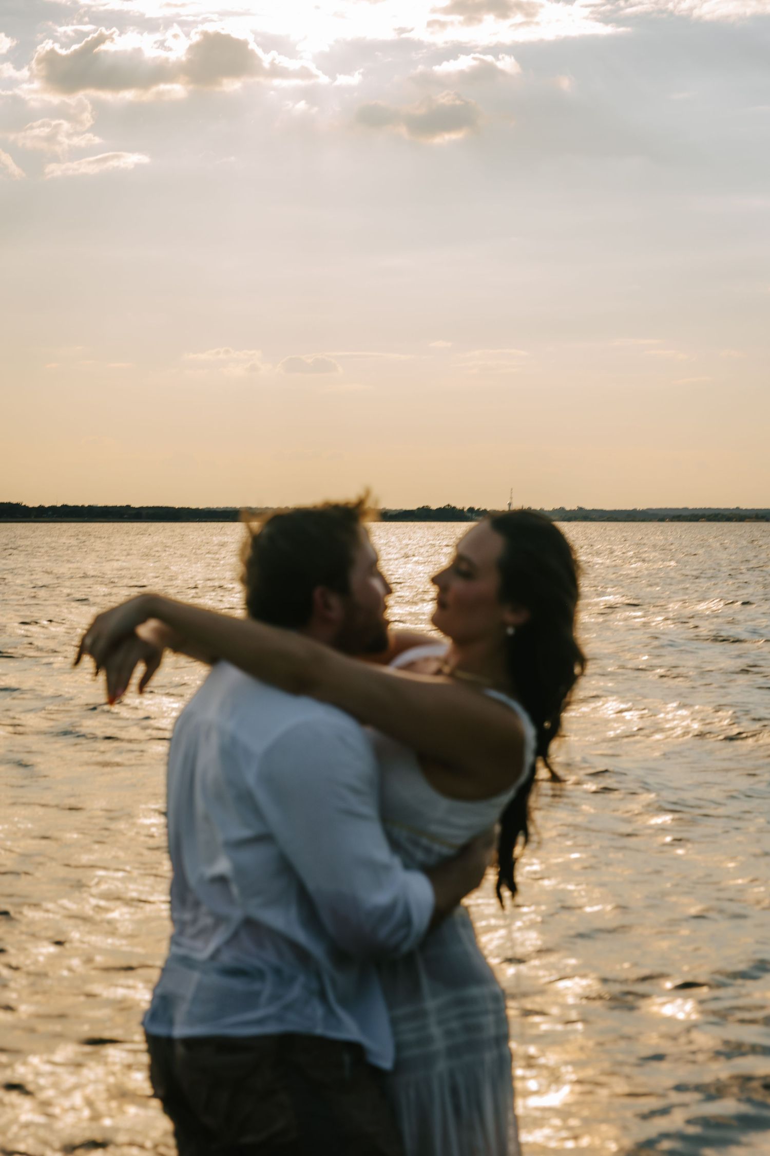 Couple embracing in shallow water at sunset, with golden light shimmering on the waves and a soft pastel sky over the horizon.
