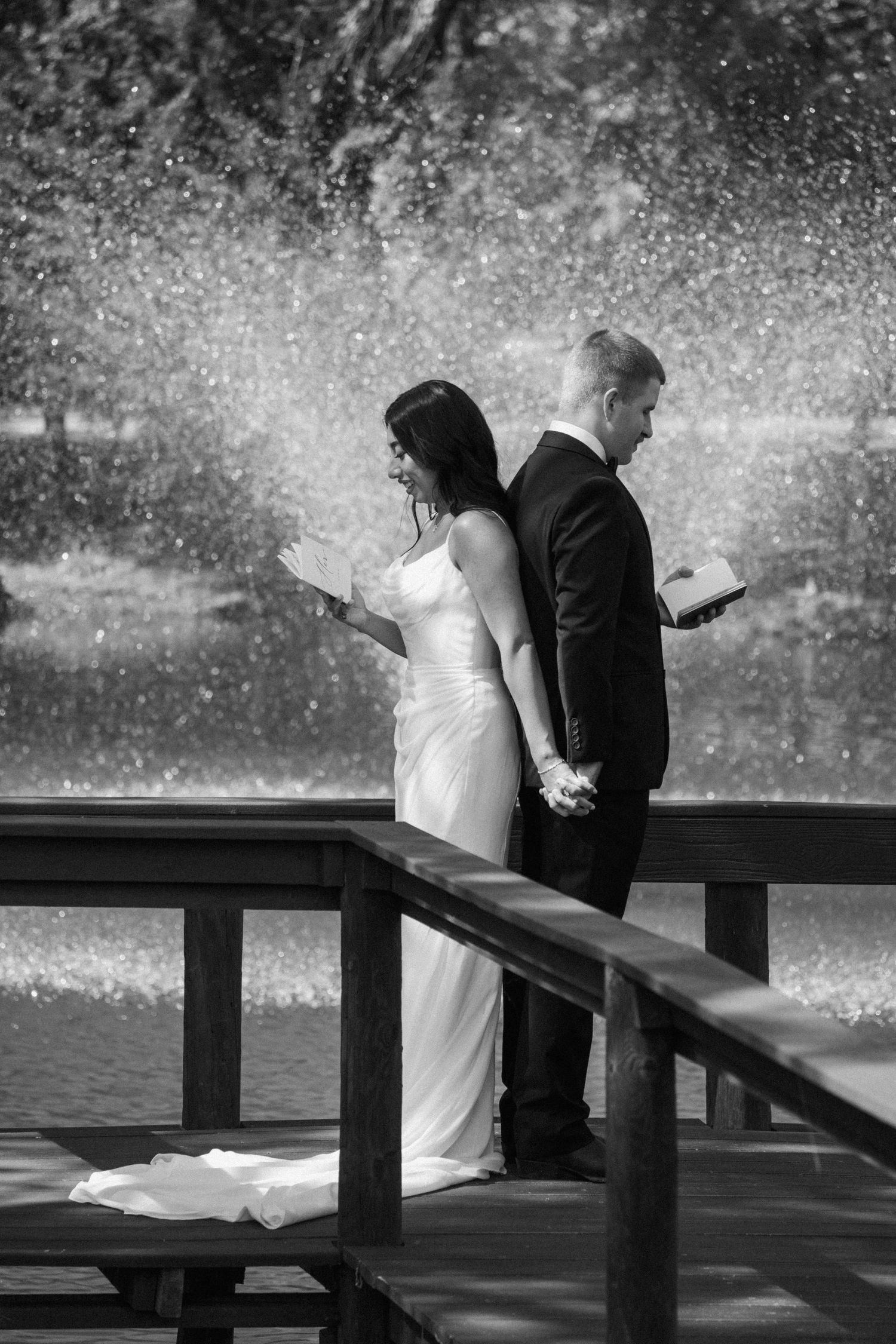 Black and white photo of bride and groom standing back-to-back on a dock, reading vows near a fountain.