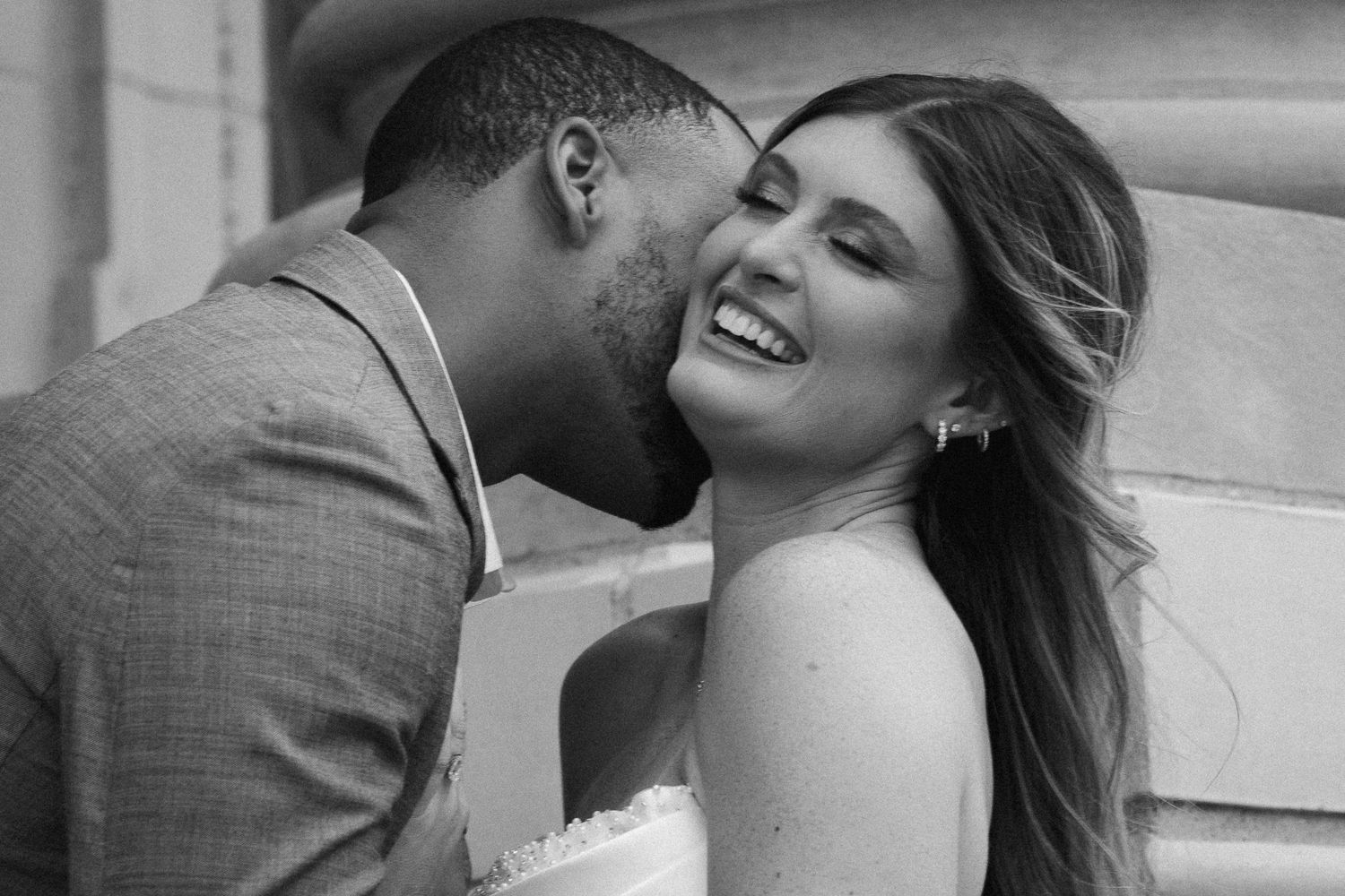 Black and white photo of a groom kissing his laughing bride on the cheek.