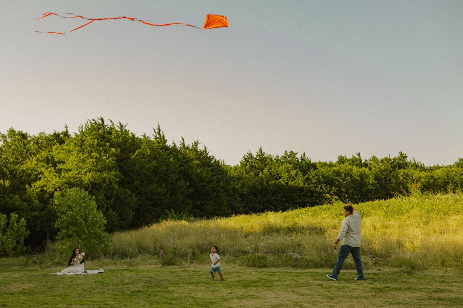 Family flying an orange kite in a green meadow with trees in the background on a clear day.