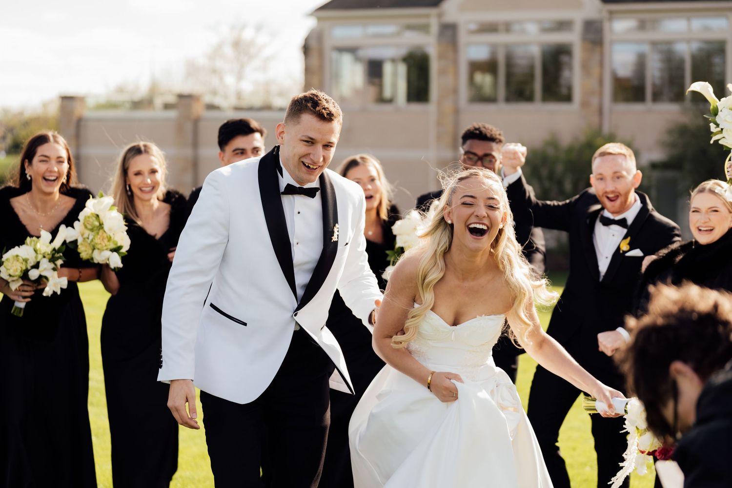 Joyful bride and groom in white tuxedo walk together outdoors, surrounded by smiling wedding party in black attire.