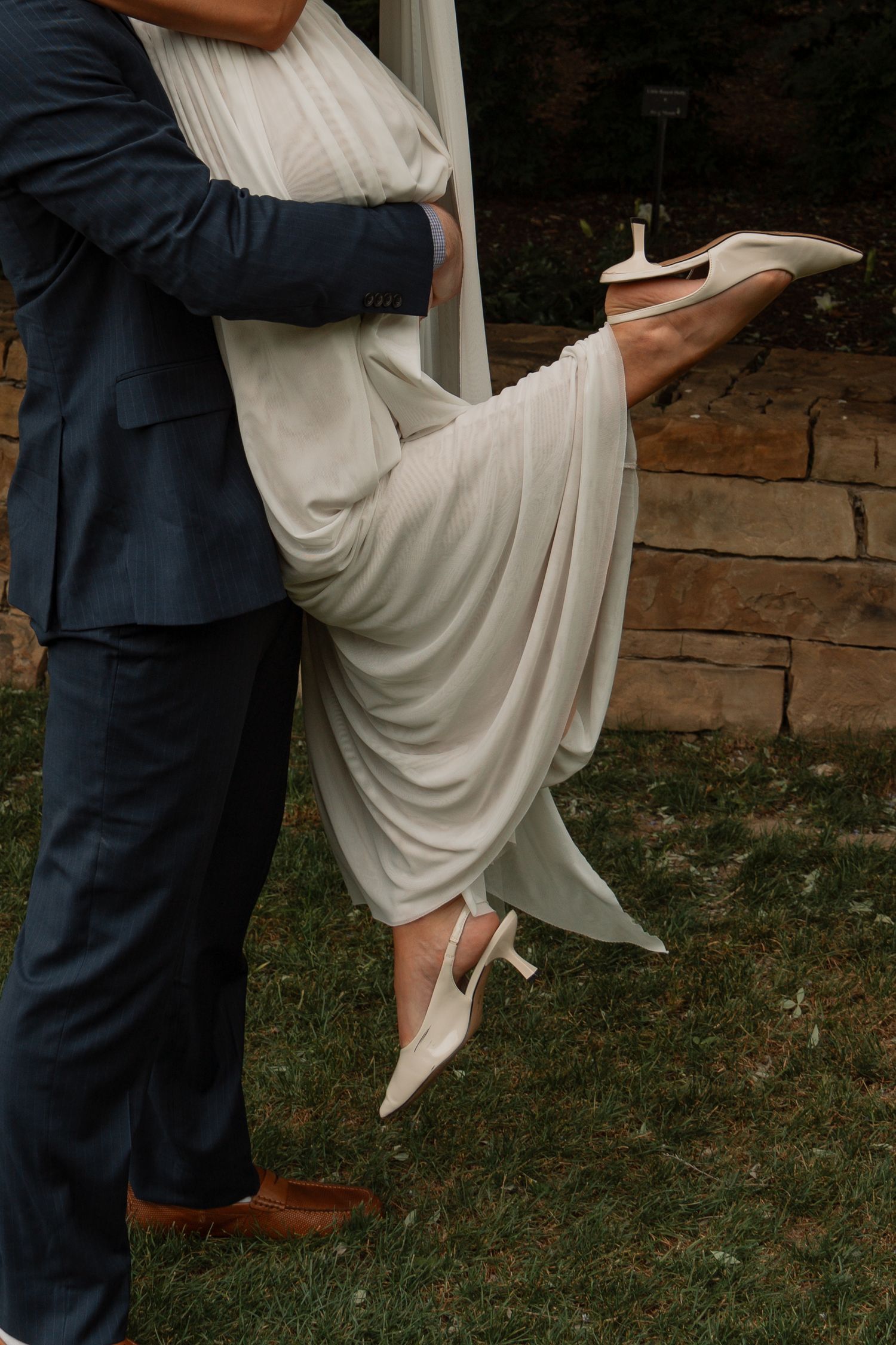 Close-up of bride in flowing white dress being lifted by groom in navy suit, showing white heels.