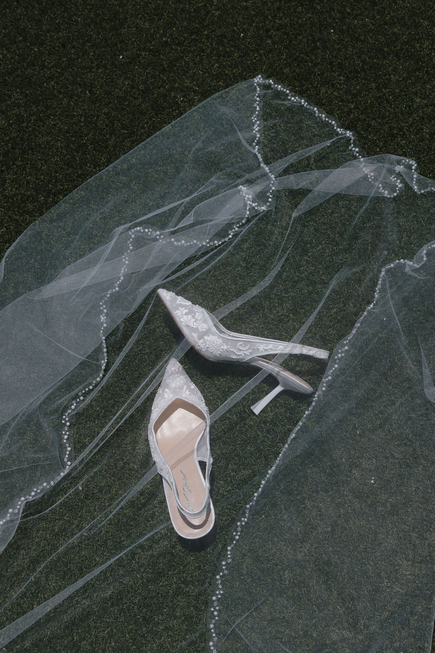 Pair of white lace pointed-toe slingback bridal heels resting on dark green grass beneath a sheer bridal veil with delicate beaded edging.