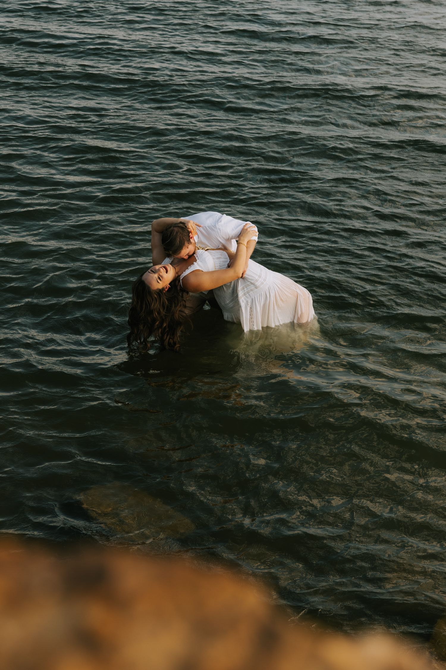 Couple in white clothing embraces in a romantic dip while standing waist-deep in dark rippling lake water at sunset.