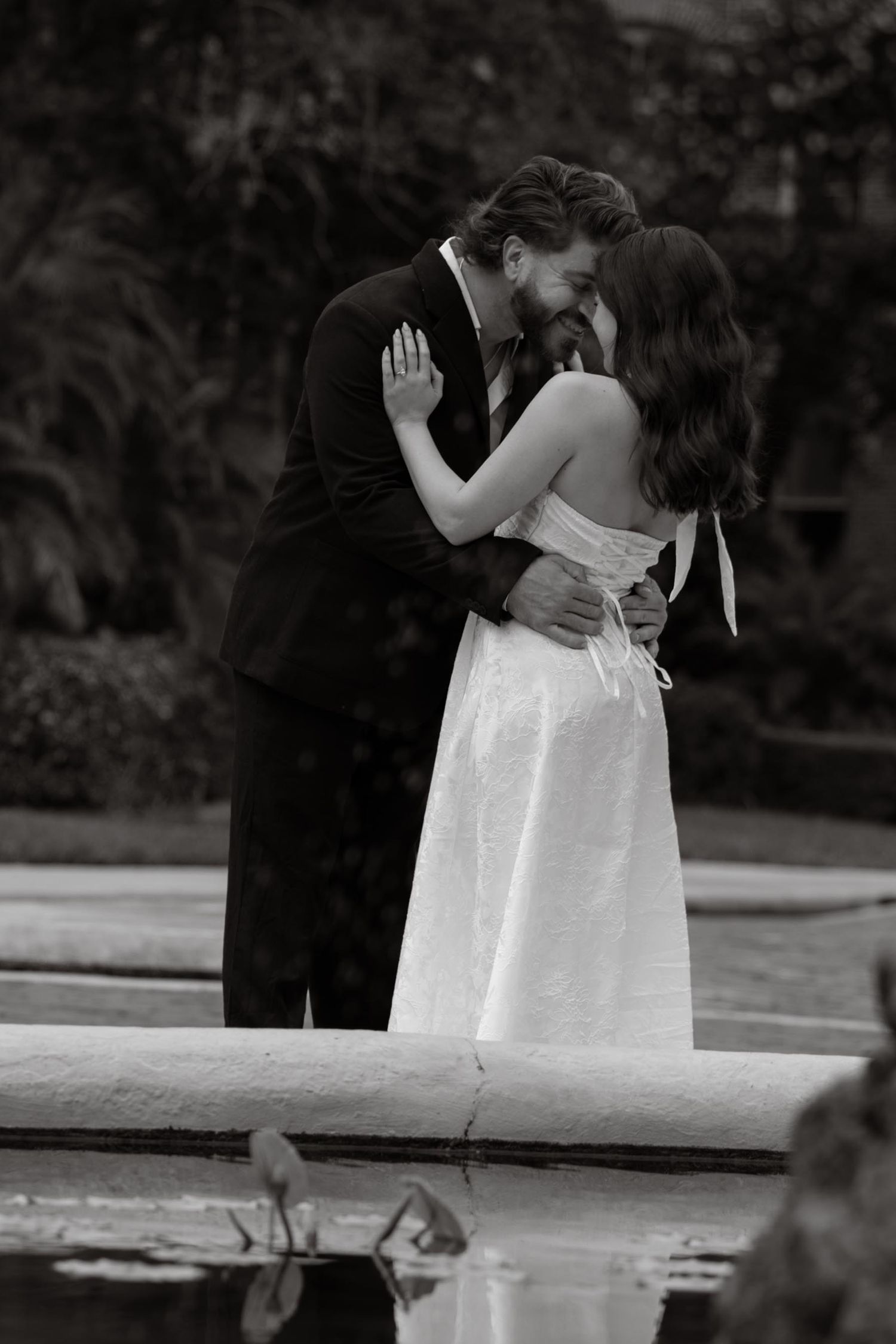 Black and white photo of a couple embracing near a garden pool, the bride in a white strapless dress and the groom in a dark suit.