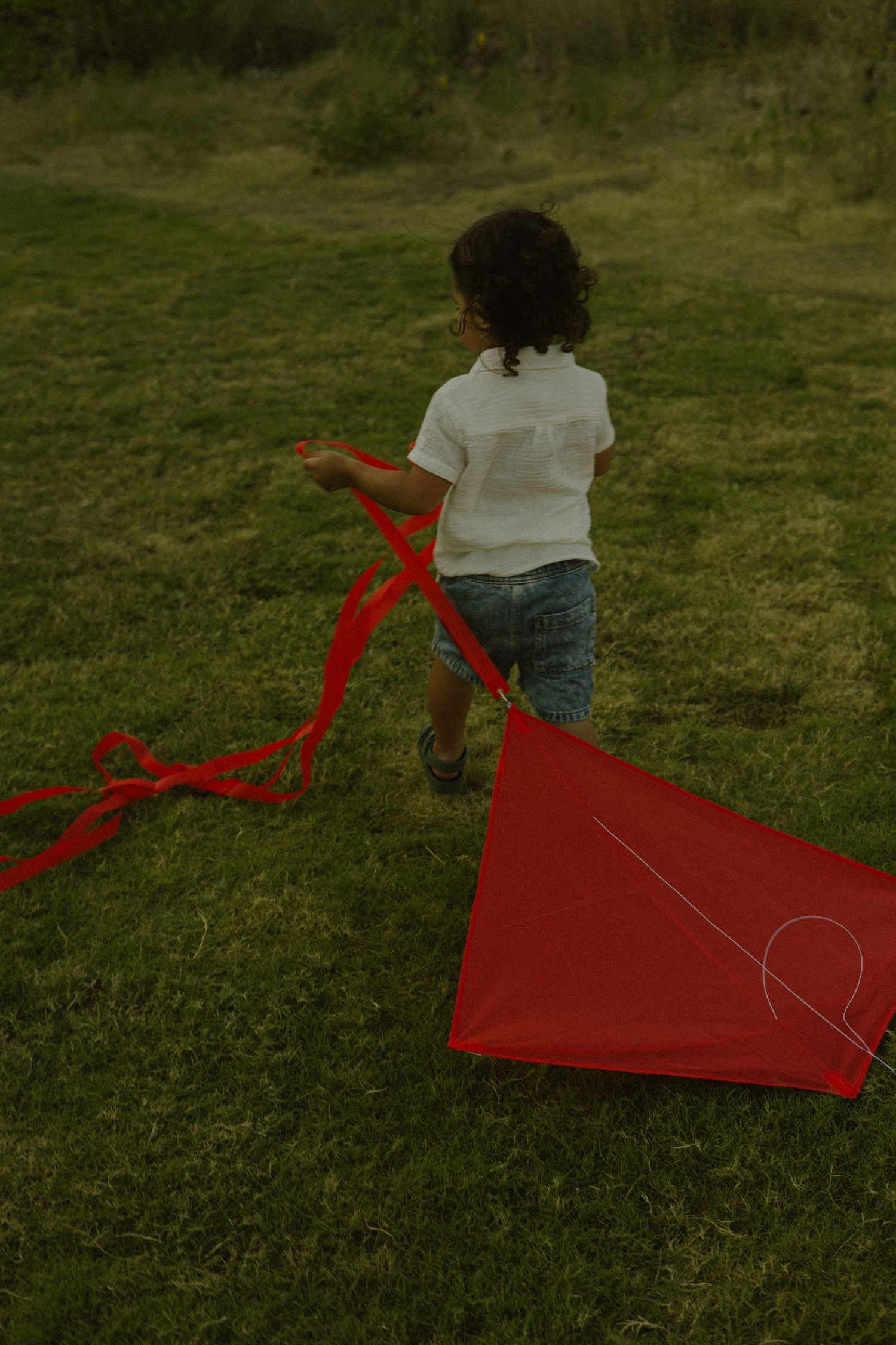 Child running on grass field with a red diamond kite with long tail streamers on a cloudy day.